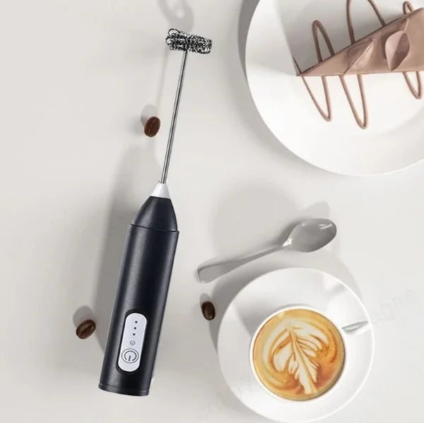 Black handheld electric milk frother placed beside a cup of coffee with latte art on a white surface, perfect for making creamy foam for lattes, cappuccinos, and other coffee drinks.