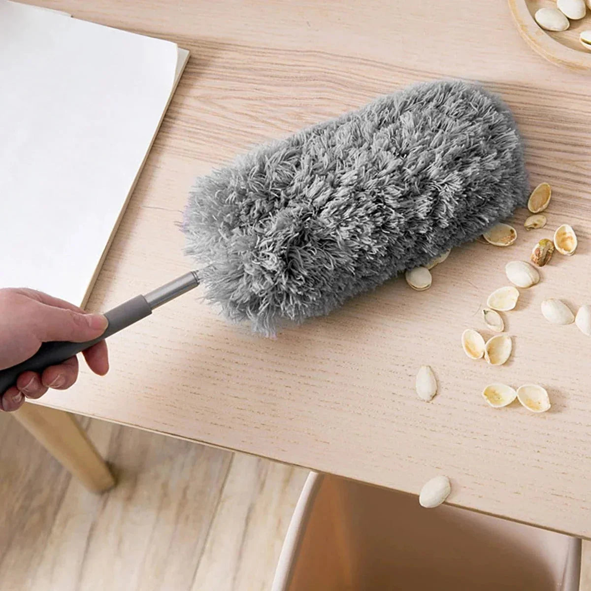 Gray fluffy dusting and cleaning tool being used on a wooden surface with scattered seeds. Demonstrates effective dust and debris removal for furniture, countertops, tables, and household surfaces, making everyday cleaning quick and convenient.