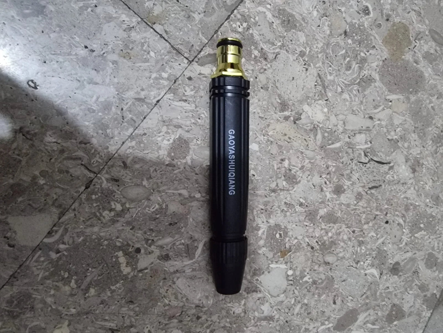 Heavy-duty metal spray nozzle attachment for a cordless pressure washer placed on a tiled floor. Durable black and brass design with adjustable spray settings for car washing, driveway cleaning, patio maintenance, and garden watering applications.