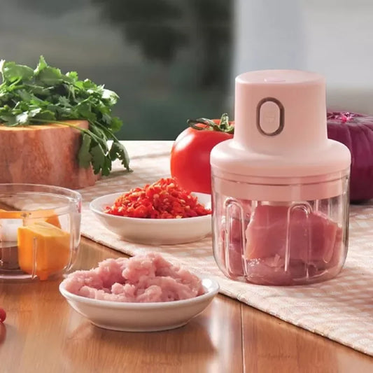 Pink manual meat grinder on a kitchen countertop with freshly ground meat, vegetables, and food preparation accessories.