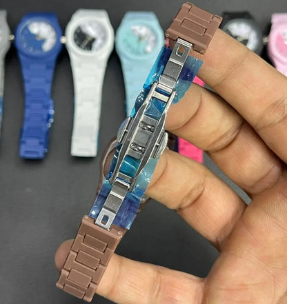 Close-up view of a stylish wristwatch showing its secure stainless steel folding clasp and durable strap design. The watch is held in hand with multiple color variants displayed in the background, highlighting comfort, durability, and everyday fashion appeal.